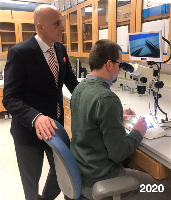 two men in a lab setting, standing man is bald with a suit and tie, the seated man has dark hair, glasses, green sweater and using a microscope and tools