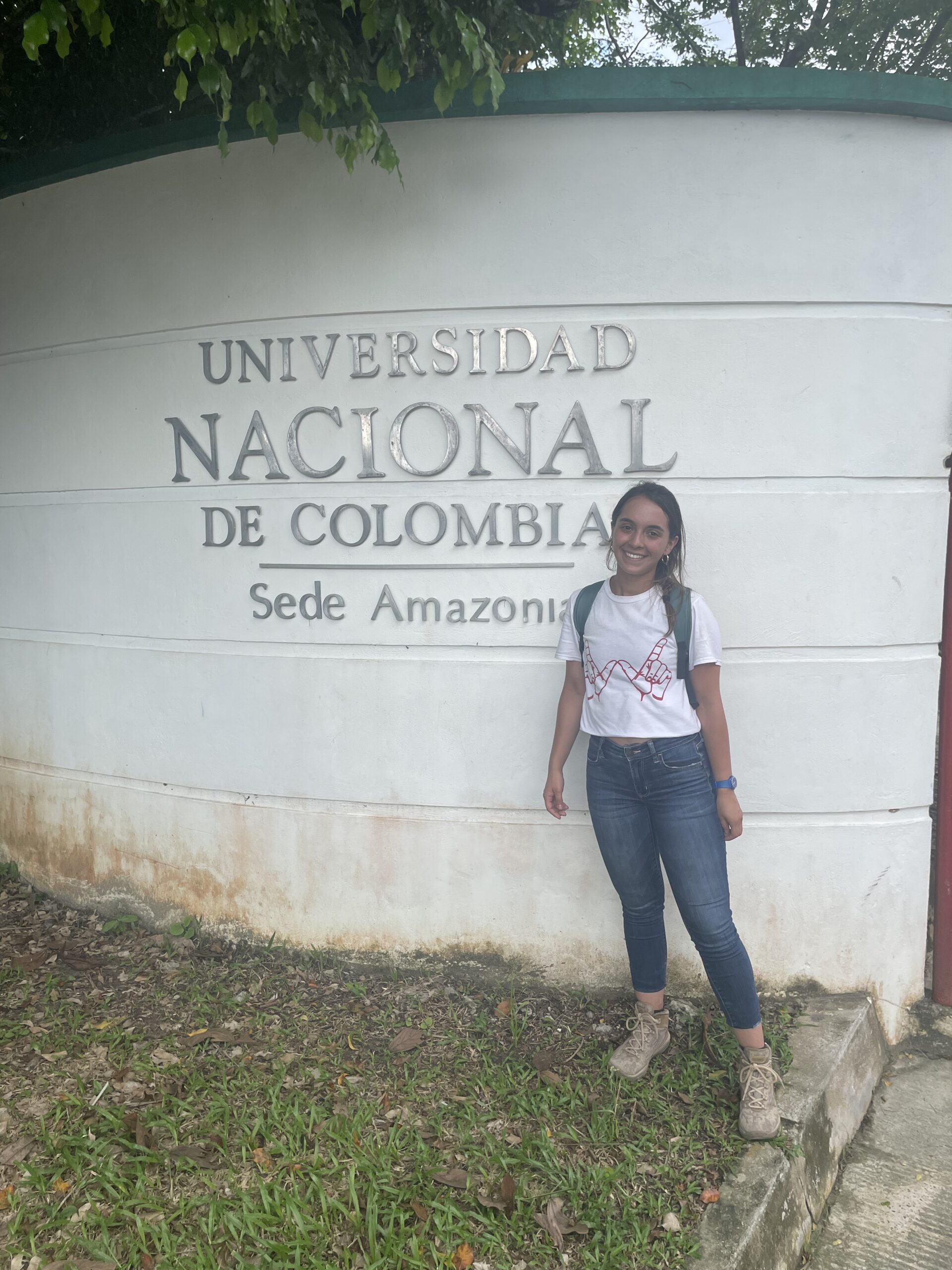 woman with dark hair, white shirt & jeans wearing a backpack in front of a universidad nacional de colombia sede amazonia sign on a building