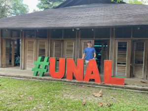 man standing behind a sign with green # and red UNAL in front of a brown building with horizontal shutters