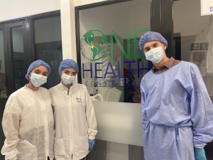 three people in scrubs and white lab coats in front of a sign with One Health Center in green & blue text