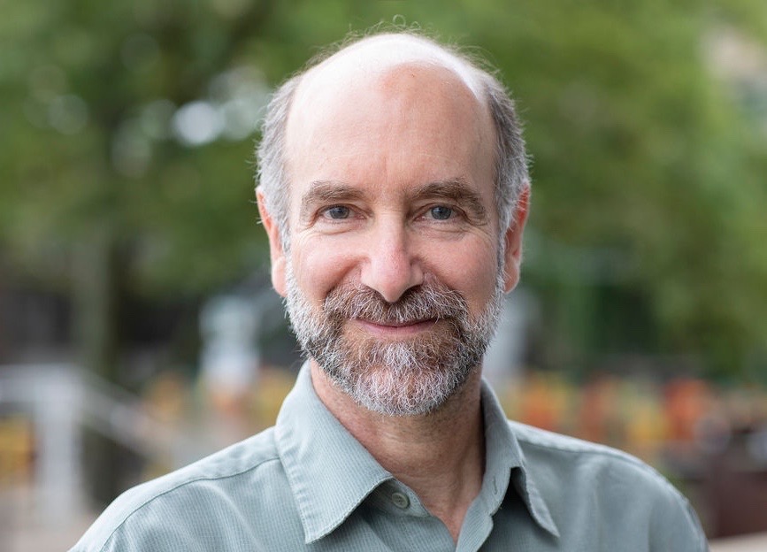 balding man with grey beard in a green shirt agains an outdoor background