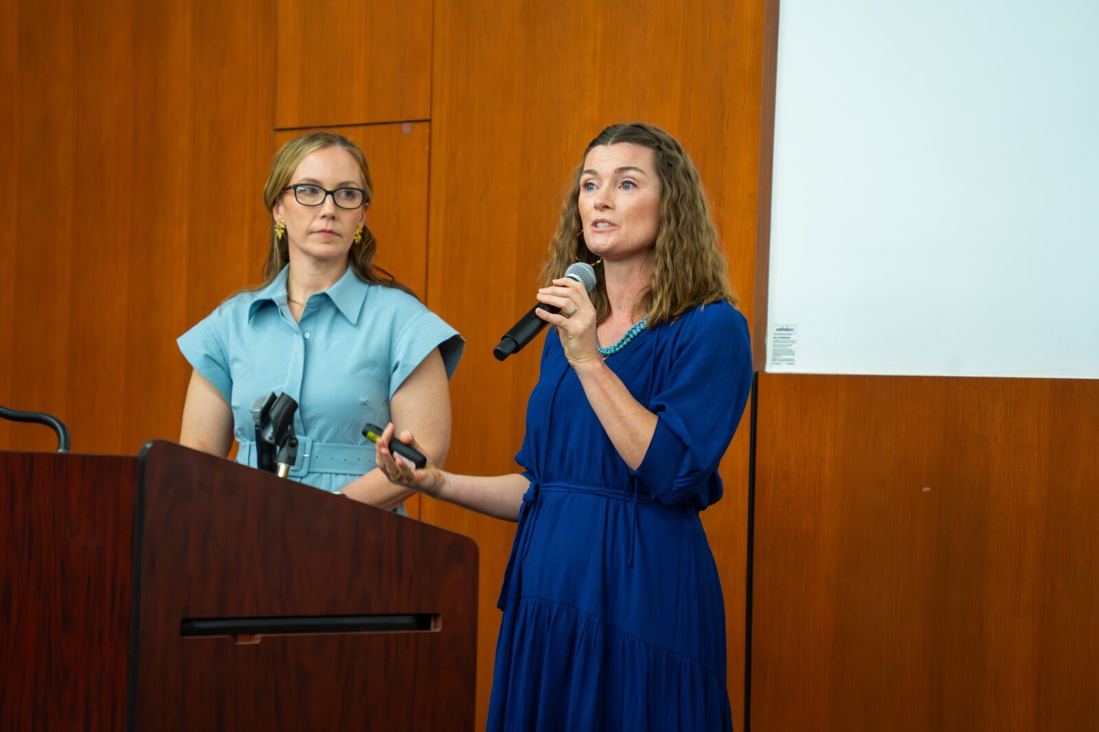 two women in blue dresses standing next to each other presenting at a lectern, woman on right is holding a microphone