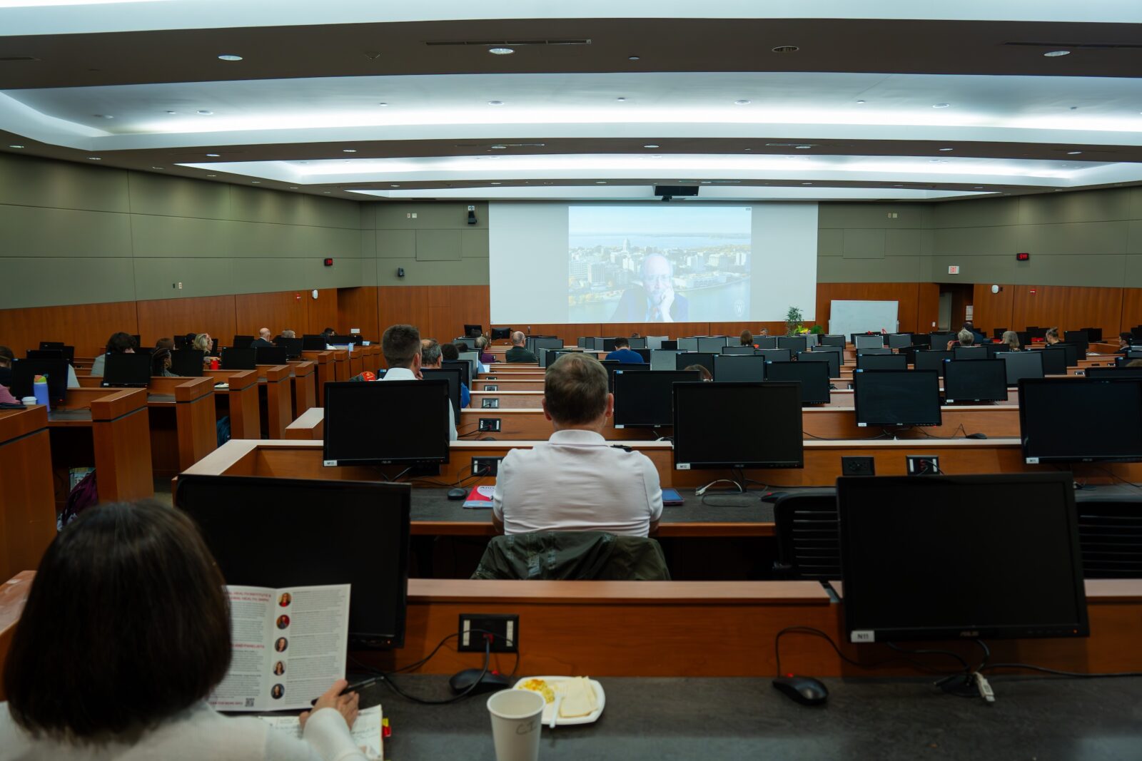 people sitting in a lecture hall looking at a screen with man holding his head in his hand