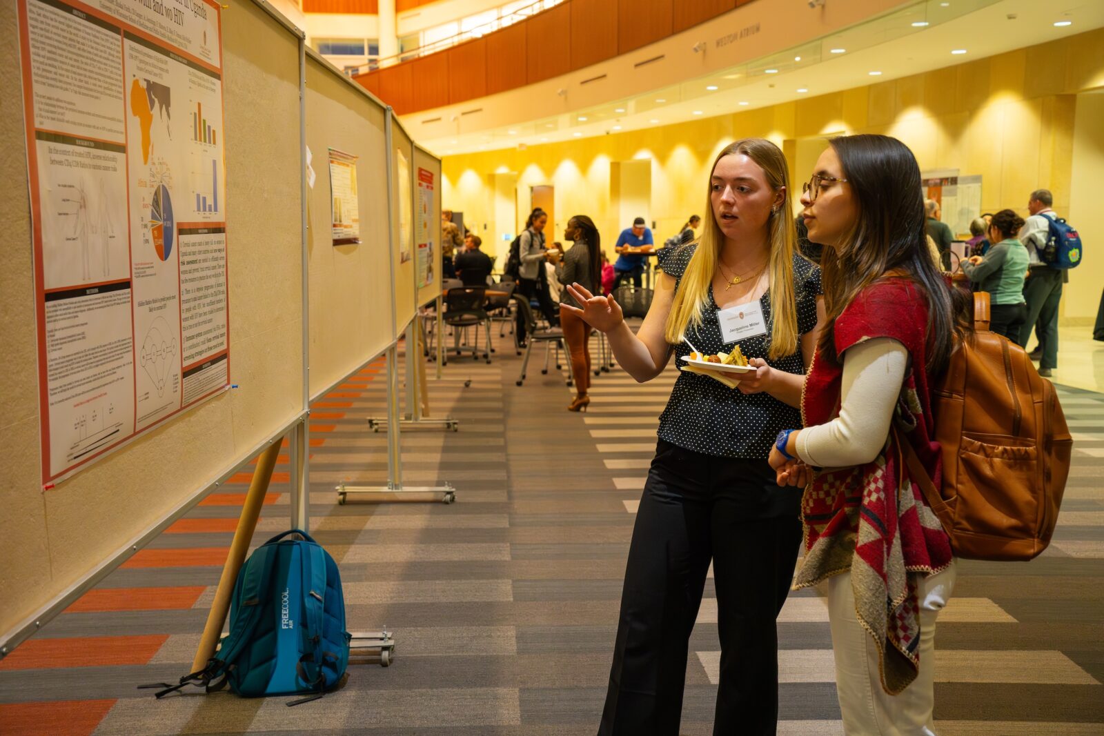 two women stand hear a poster, one is holding a plate of food, the other is wearing a backpack