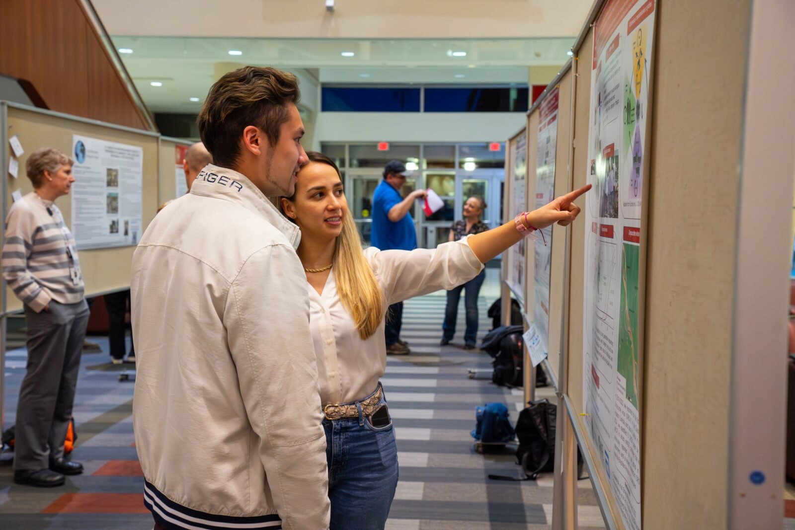 a woman with light hair and white shirt points at a poster with a man with dark and light hair looking at the poster