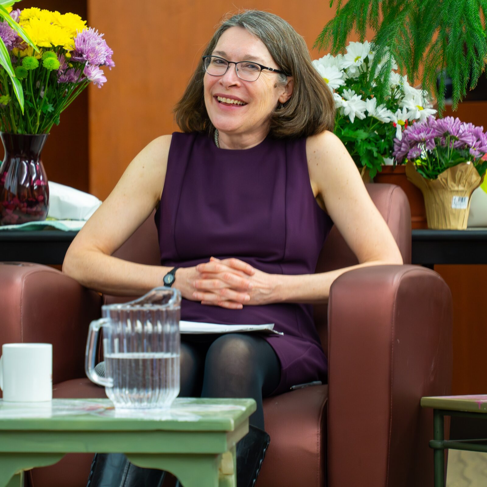 woman in purple dress sitting with hands folded with flowers in thehbackground