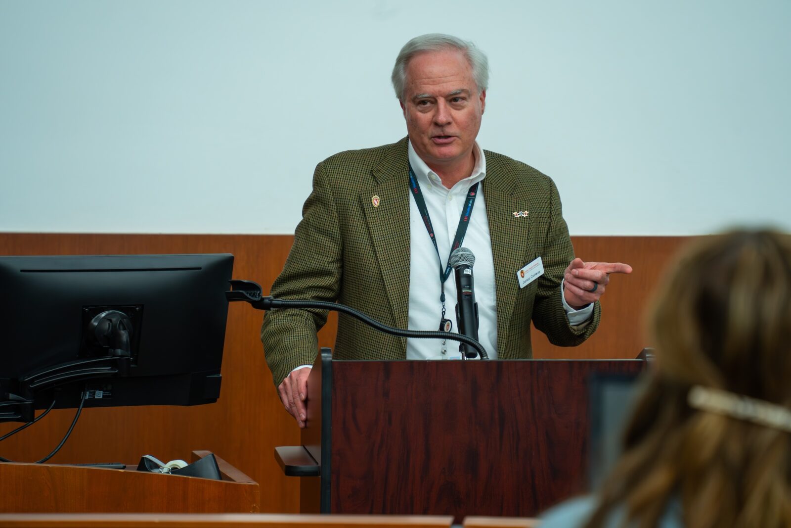 man with white hair at lectern wearing a green jacket and white shirt speaking with a microphone and pointing his finger