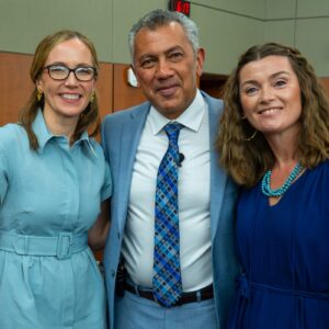 two fwomen in blue dresses with one man in a blue suit with white shirt in a lecture hall
