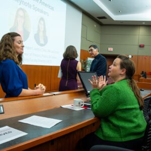 two women chatting, woman on left standing and wearing a blue top, woman on right sitting and wearing a green top, a screen wth two photos in the background along with two other people talking
