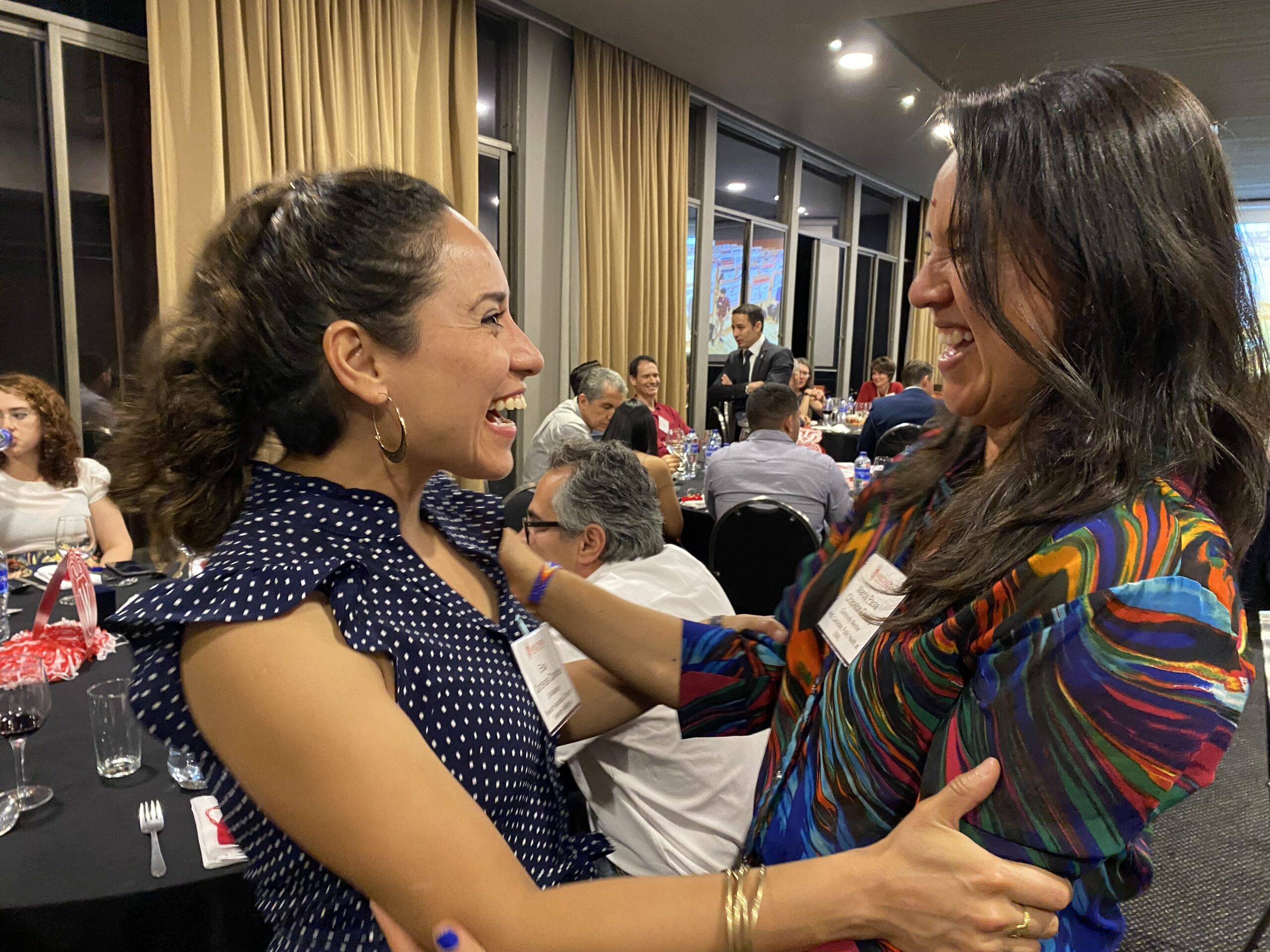 two women greeting each other with smiles and hugs in a room of people sitting