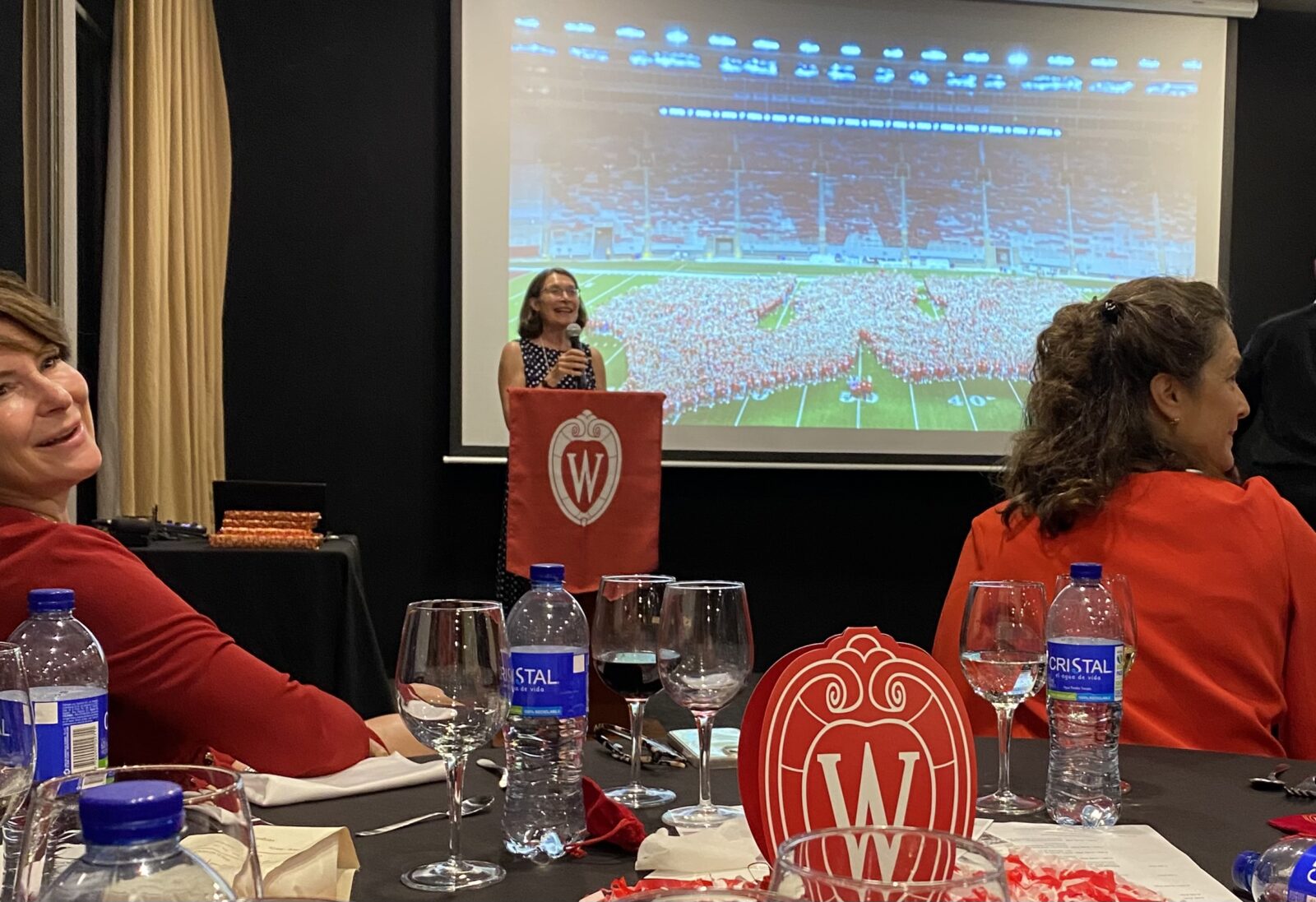 UW Madison Vice Provost and International Division Dean Frances Vavrus presenting at the Badger Salud event, Sept 2023; woman at lecturn wth uw rest and two women at table watching
