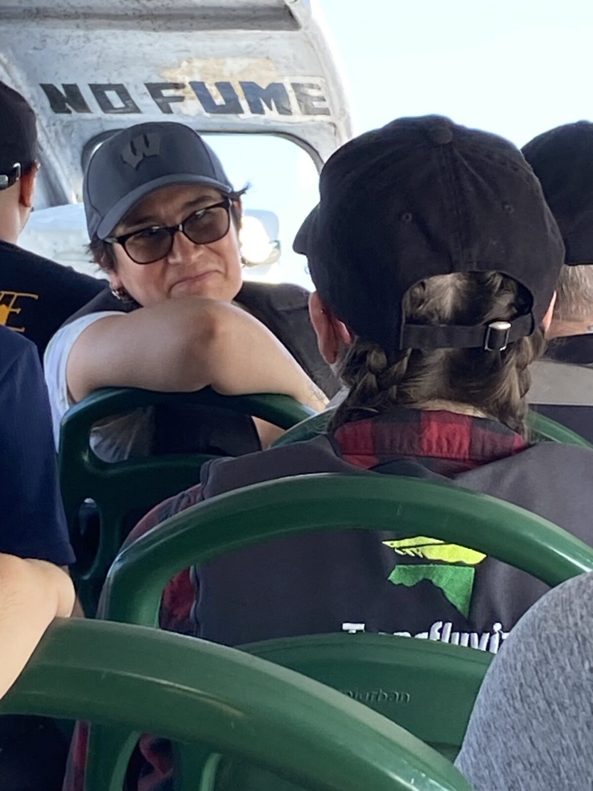 GHI Post Doc Olga Lucia Chaparro sharing information on the Amazon River, Sept 2023; woman with a black hat & sunglasses in a boat with a no fume sign behind her and another woman with braids and a hat looking at her