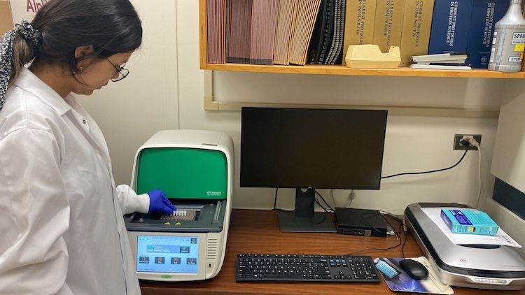 woman with dark hair and glasses in a white lab coat using lab equipment on a desk with a computer monitor