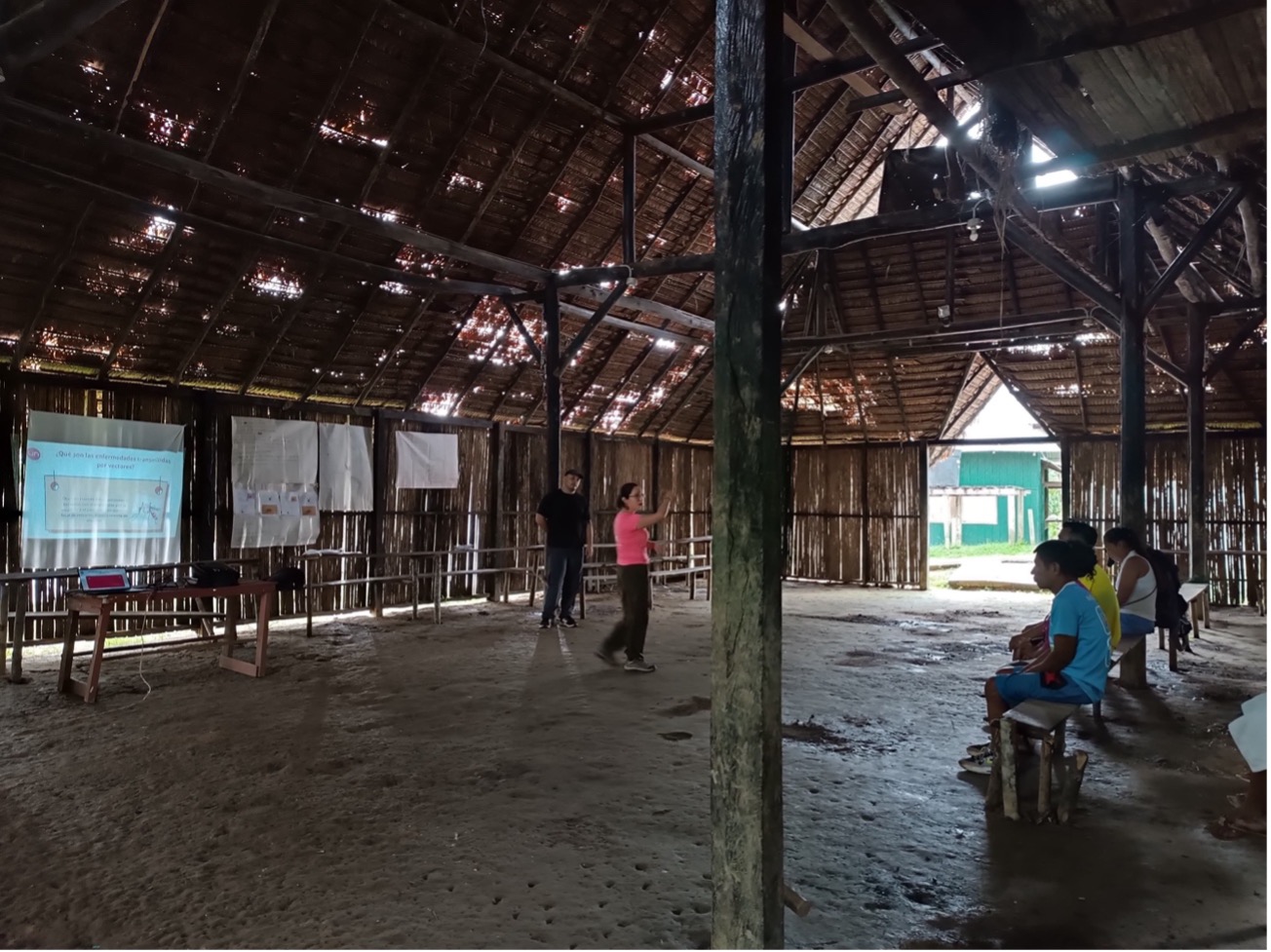 June 2024, two people standing under a wood roof of a building with other people sitting and a screen on the back wall