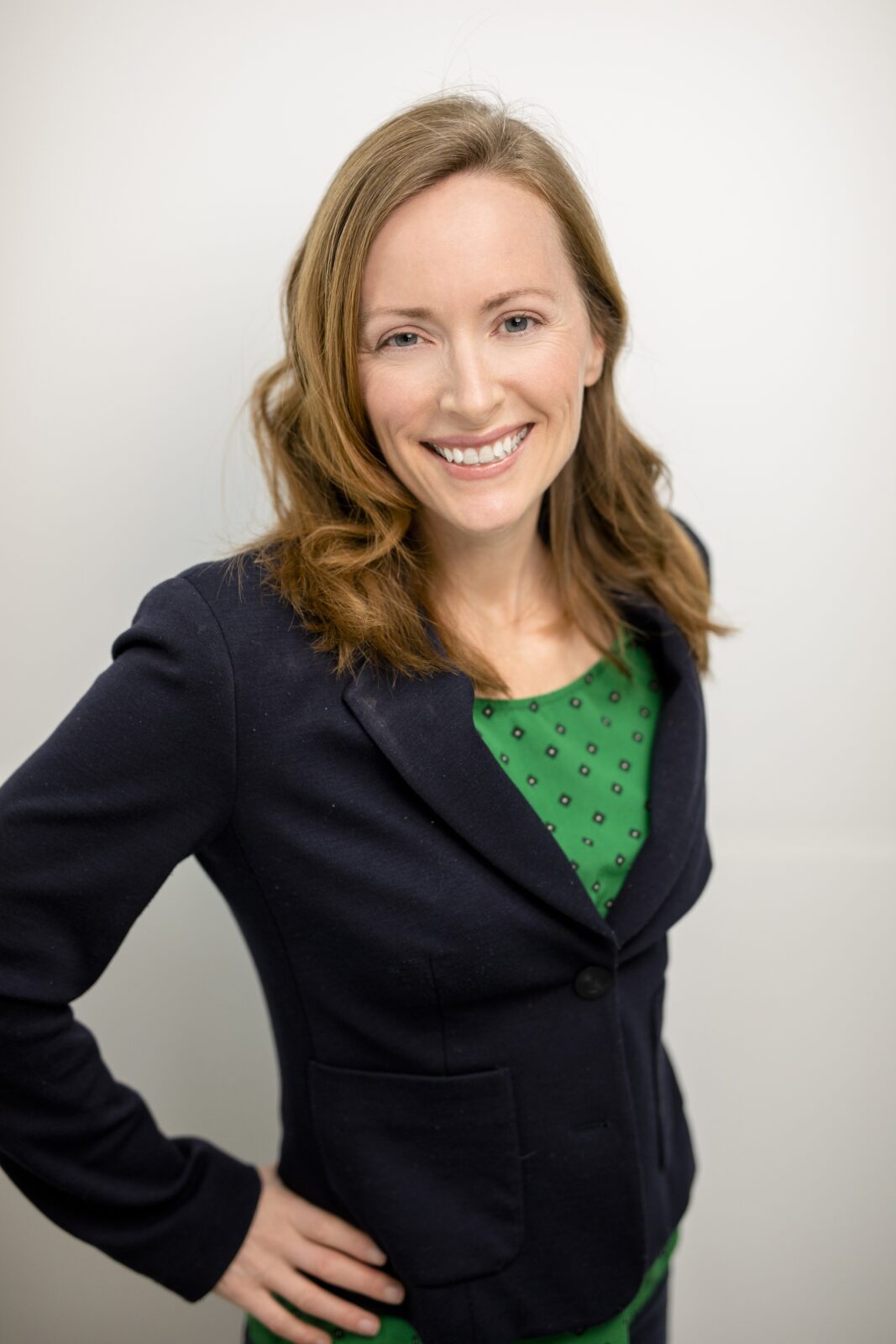 woman with light hair, blue jacket, gren and blue polka dot shirt against a light background