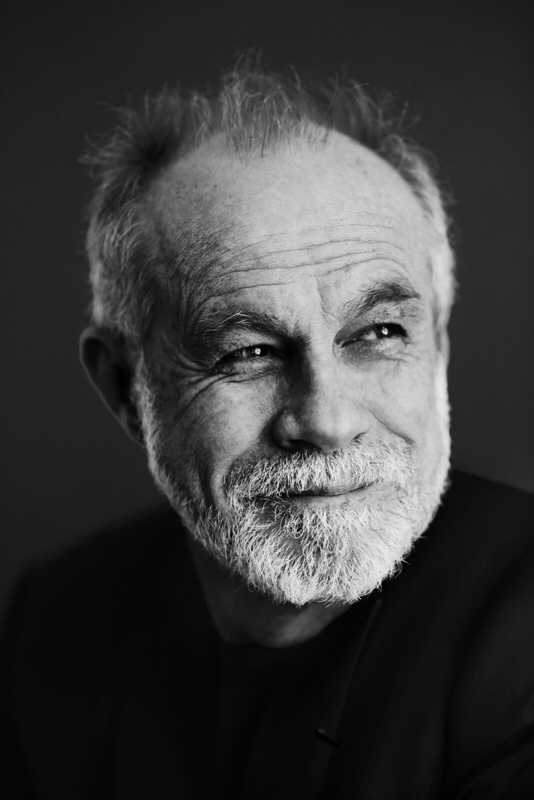 black and white photo of a balding man with white beard in a dark top agains a dark backgroun