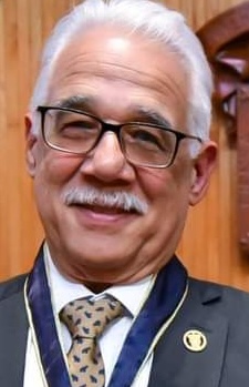 man with white hair & mustache, glasses, dark suit with white shirt and paisley tie against a brown background