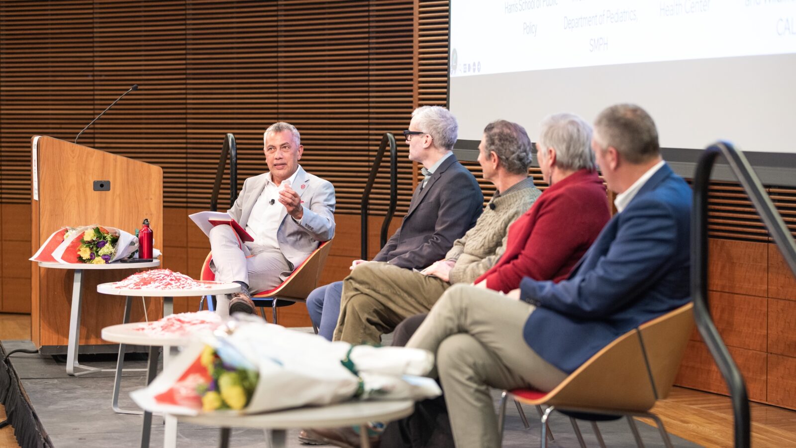 five people sitting in chairs on a riser with a lectern and screen behind them