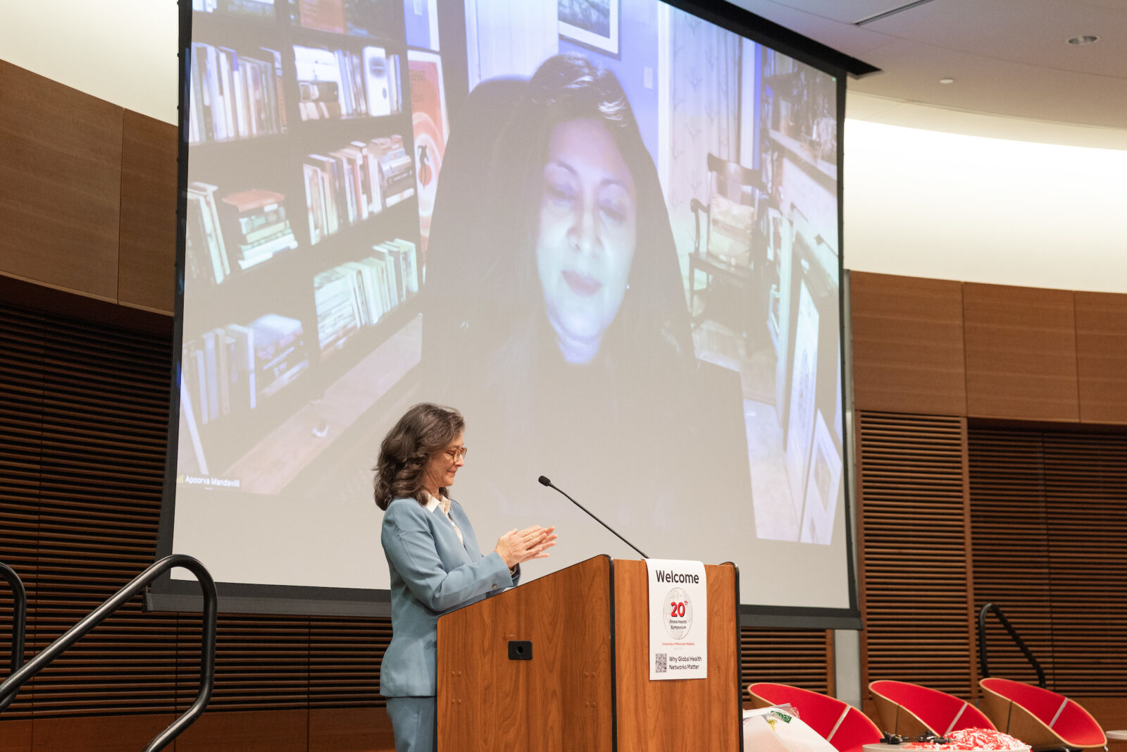 a woman at a lectern clapping hands as another woman with dark hair is on the screen in the background