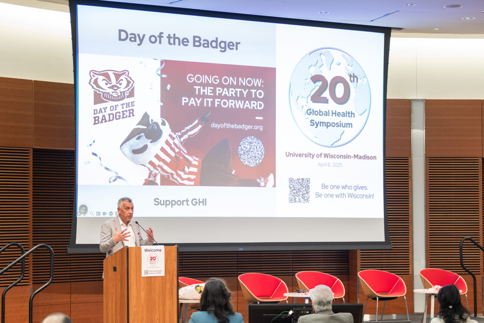 a man at a lectern with a screen featuring Bucky Badger and a 20th Global Health Symposium logo in the background