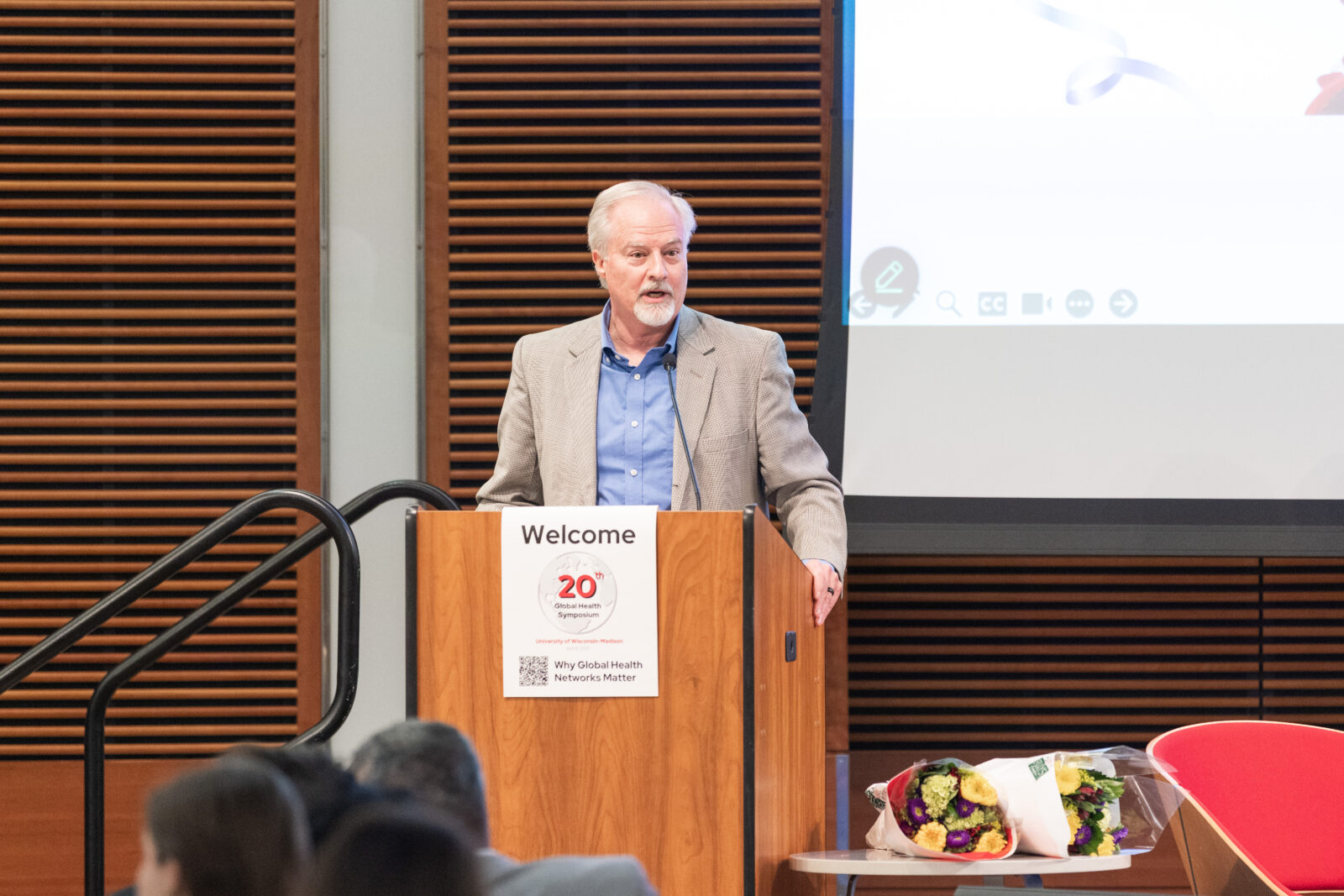 man with white hair, light jacket, blue shirt at a lectern with a screen behind