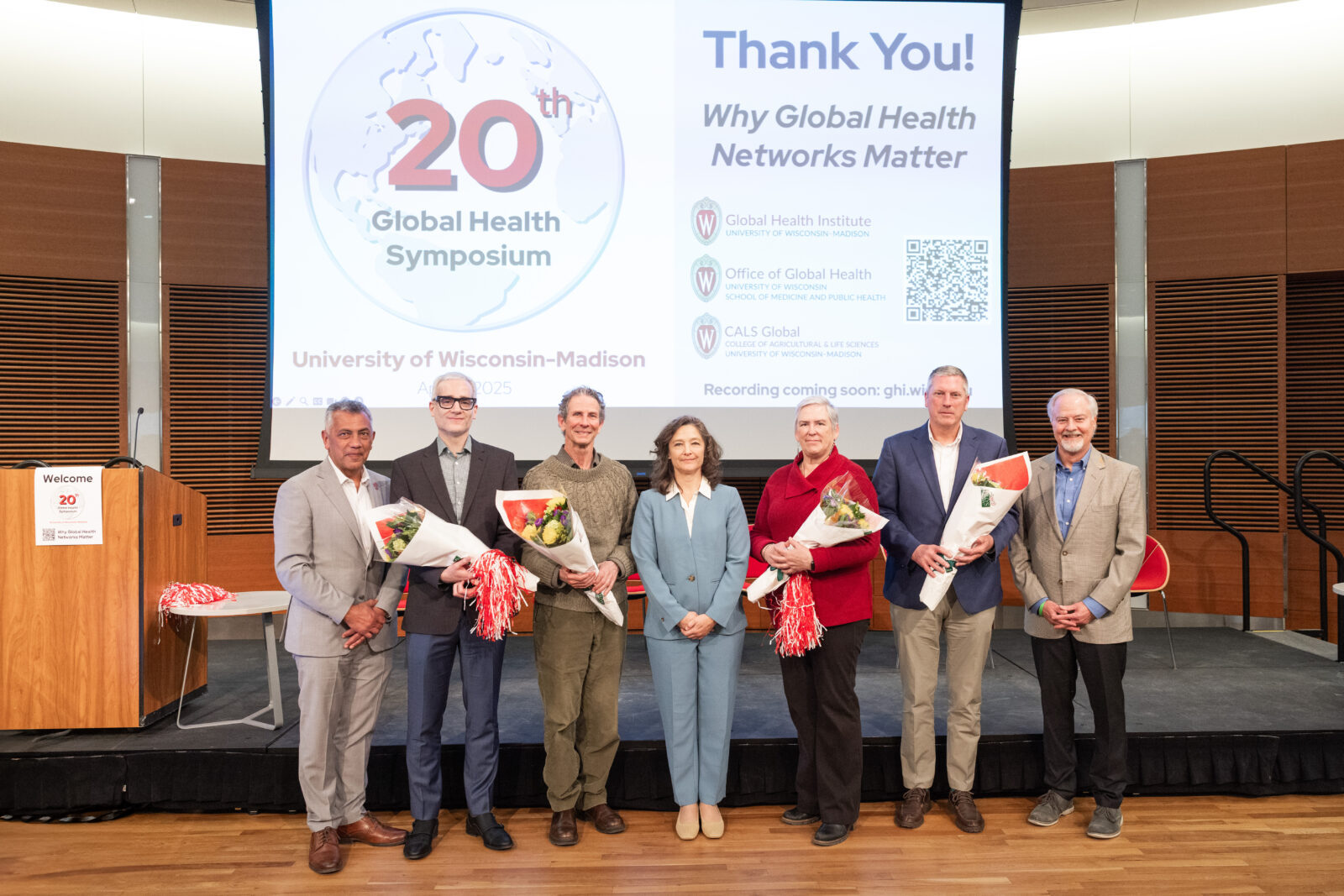 seven people standing in front of a screen with the 20th global health symposium logo