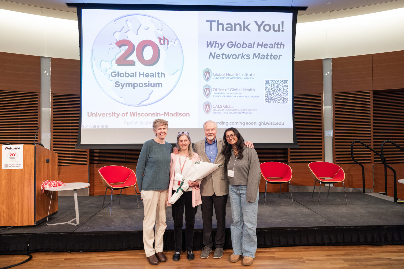 four people standing with the 20th global health symposium logo on a screen in the background