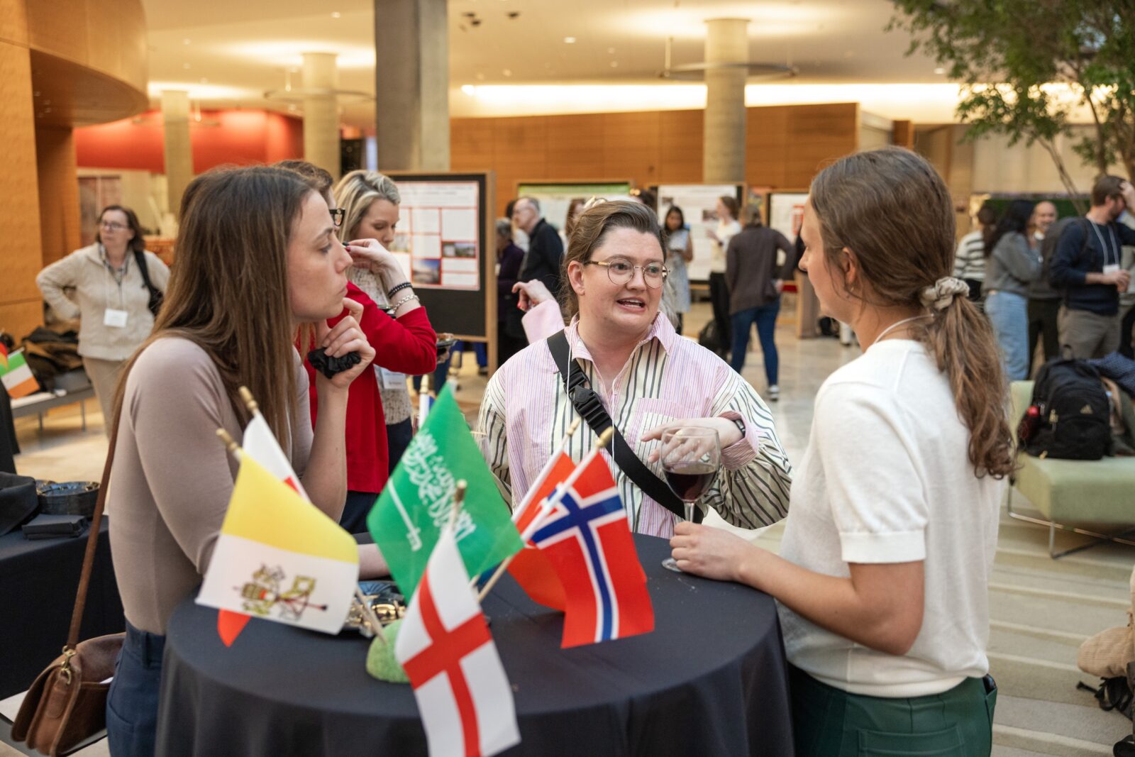 three women talking and smiling while standing at a table and others looking at posters in the background