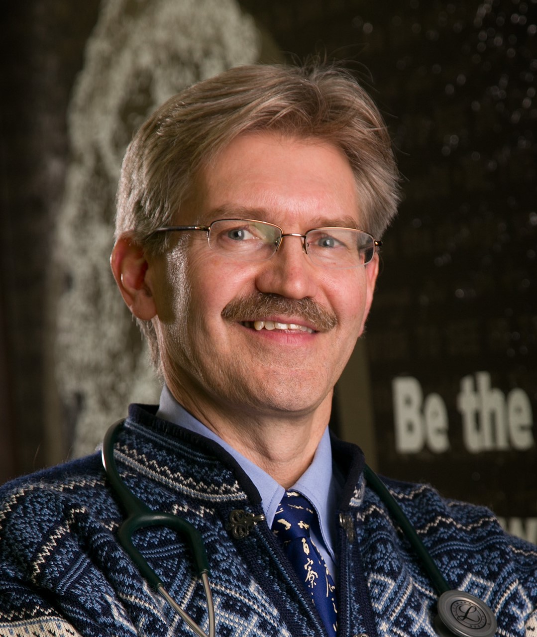 headshot of Jonathan Temte, man with light brown hair, blue, white & black sweater with blue shirt & tie against a grey background with the words Be the in the background