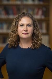headshot of Jennifer Nuzzo, woman with brown curly hair, blue top, necklace, against a background with books on bookshelves