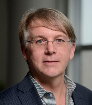 headshot of William Moss, man with light brown hair, glasses, blue jacket with striped shirt against a background of a grey room with a window