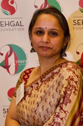 Woman with greying hair wearing a read, cream, and gold sari, against a background with the Sehgal Foundation logos