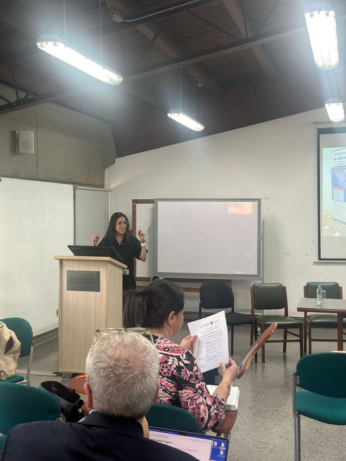 May 2024, woman at lectern with a large white board and chairs nearby with people sitting