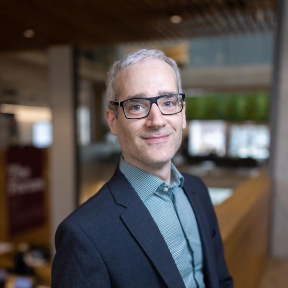 February 2025, photo of man with grey hair, & glasses, dark jacket, blue-green shirt against a room with windows and pillar background