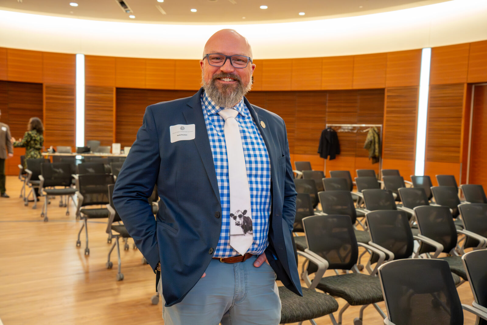 Keith Poulsen standing in front of empty chairs before the 2026 Global Health Symposium begins; man with beard wearing a dark jacket, plaid shirt, jeans, and a tie with a cow graphic