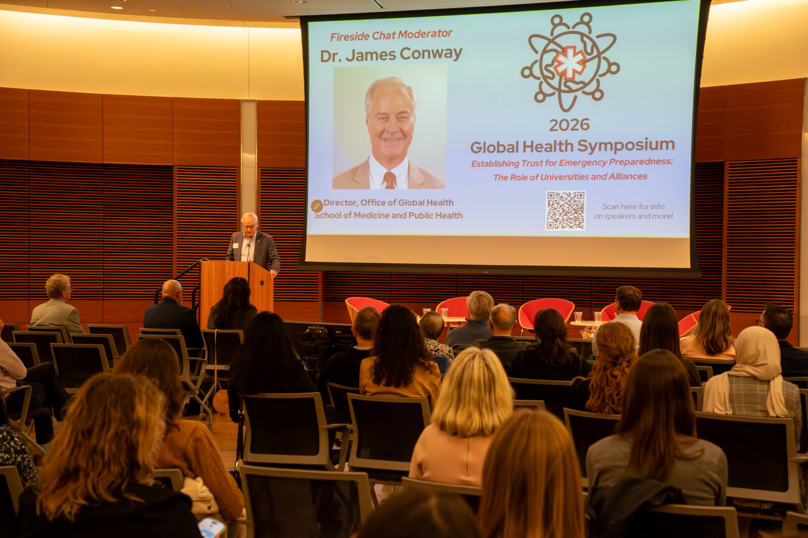 Photo from 2026 Global Health Symposium at the Discovery Building;Jim Conway at the lectern with his image and text on the screen behind him and speaking to a crowd sitting