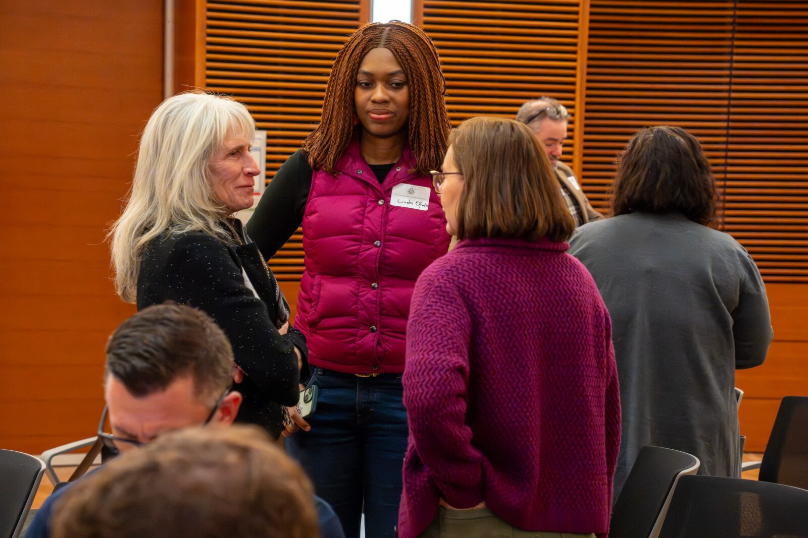 Photo from 2026 Global Health Symposium; three women standing and chatting, two wearing pink, two people standing in background and some sitting in foreground