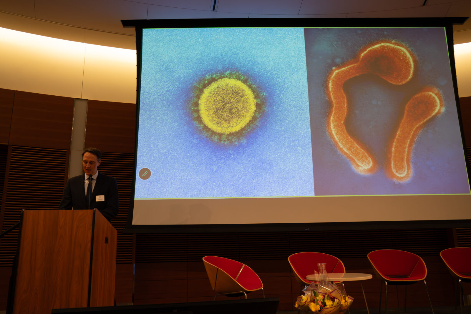Photo from 2026 Global Health Symposium at the Discovery Building; Dr Westergaard at lectern with slide on screen featuring one side with a blue background and yellow circle, the other side dark background with two orange lines