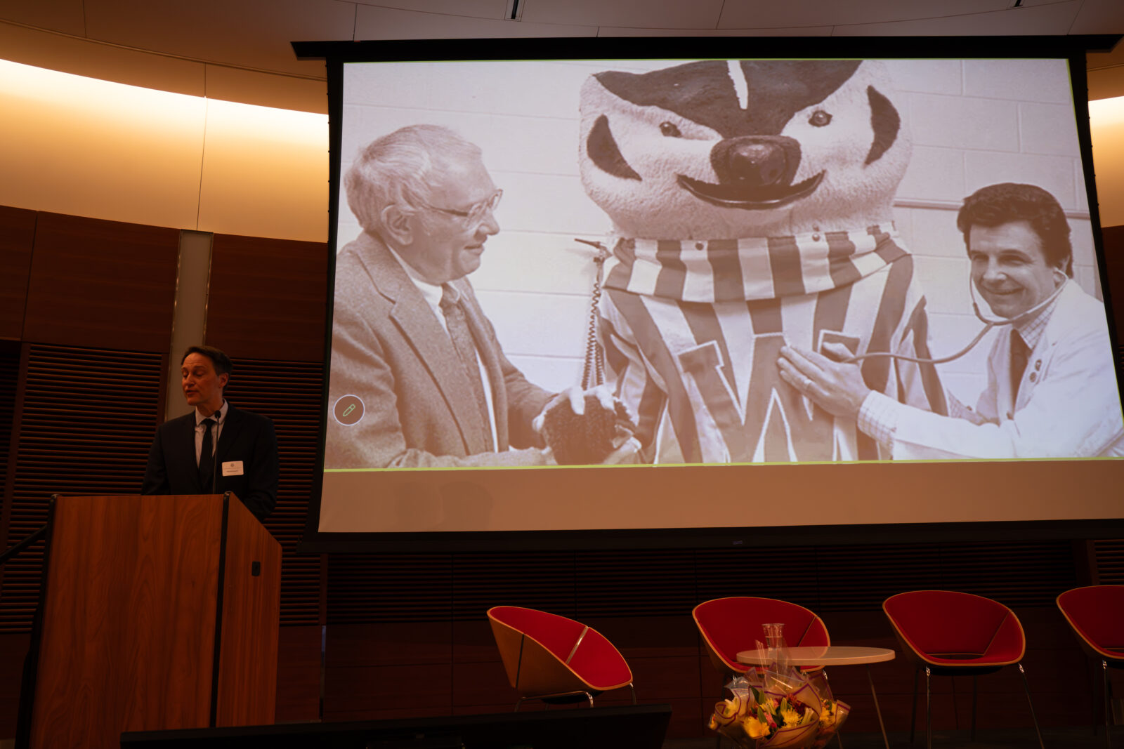 Photo from 2026 Global Health Symposium at the Discovery Building; Dr Westergaard at lectern with screen in background with a black and white photo of Bucky Badger with two men, one using a stethoscope on Bucky