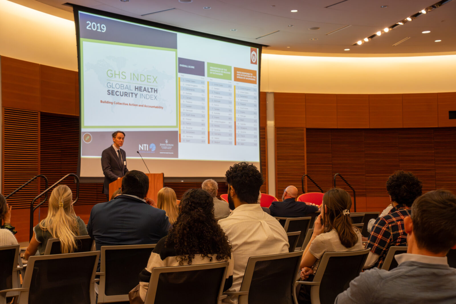 Photo from 2026 Global Health Symposium at the Discovery Building; Dr Westergaard at lectern with the audience looking on; screen is up in the background with four columns of text
