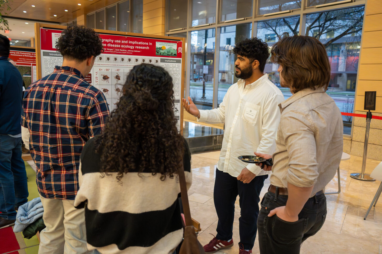 Photo from 2026 Global Health Symposium at the Discovery Building; four people standing in front of a poster, one man in a white shirt and dark hair has hand raised and speaking