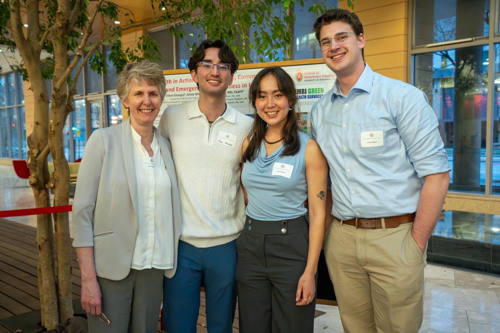 Photo from 2026 Global Health Symposium at the Discovery Building; four people (2 men, 2 women-one is Ann Evensen from OGH, SMPH) standing in front of a poster