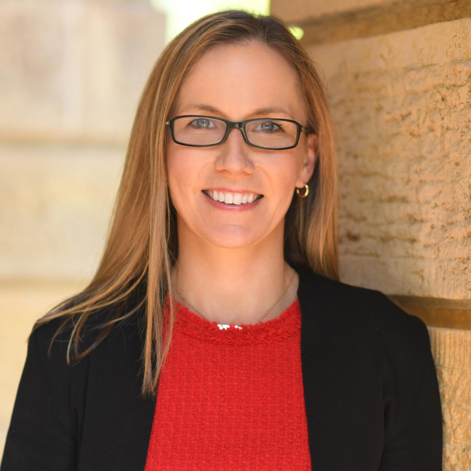 February 2024, headshot of Erin Barbato, woman with blonde hair, glasses, black jacket, read shirt against a background outside against a stone building