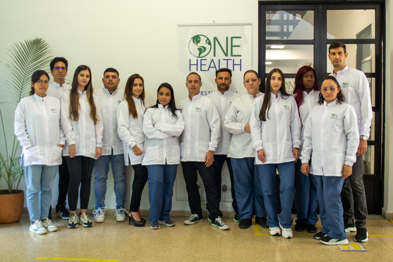 August 2024, people in white lab coats in front of a sign with green and blue letters saying one health