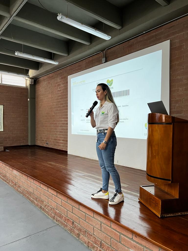 May 2024, woman with light hair, white shirt, and jeans standing on a stage with a screen holding a microphone