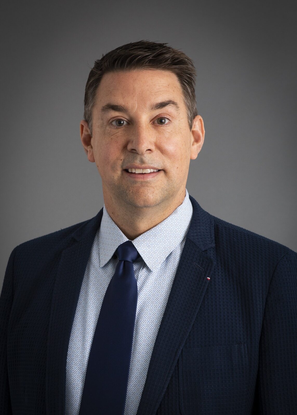 July 2024, man with dark hair, dark jacket and tie, light shirt against a dark grey background