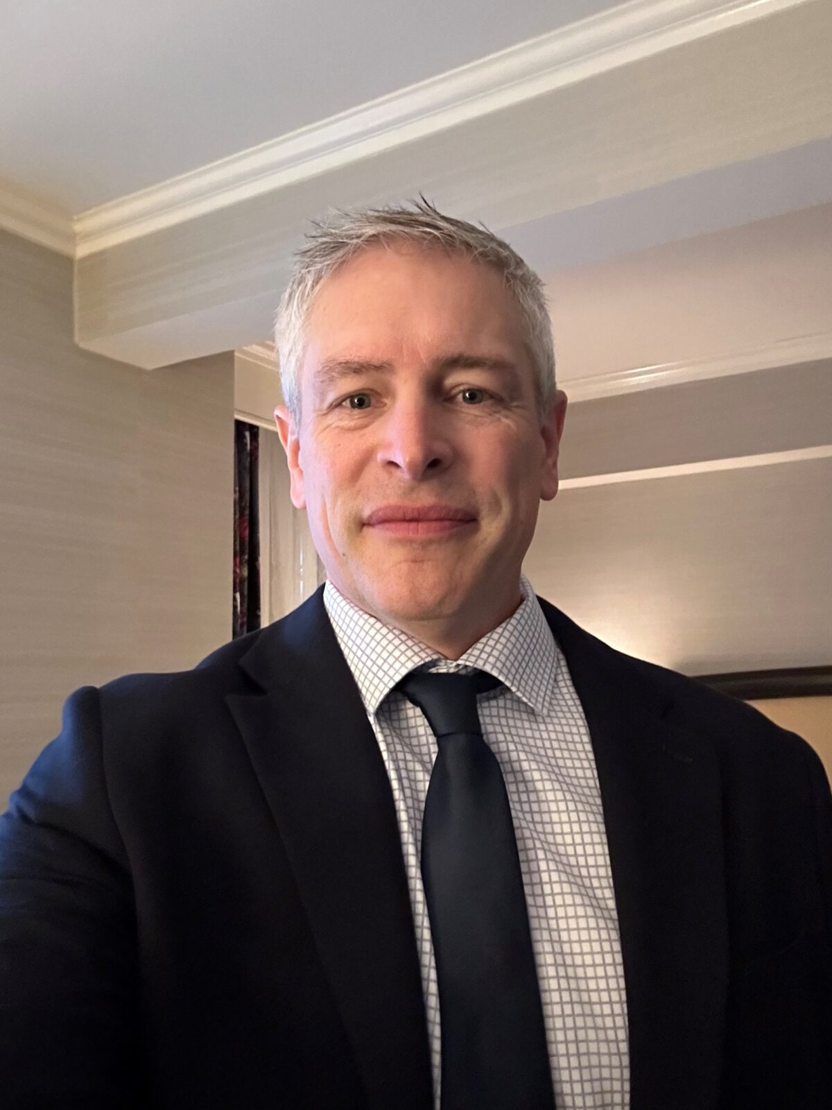December 2024, man with grey hair, dark suit wth white shirt and dark tie in a room with white3walls and ceiling

