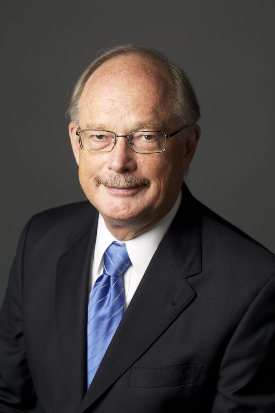 July 2023, balding man with glasses, dark jacket, white shirt, blue tie against a dark grey background