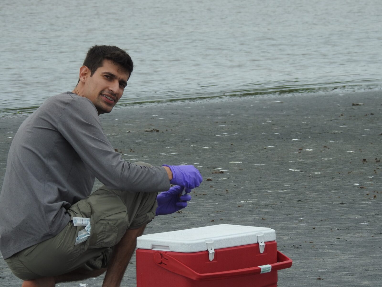January 2024 From Treating to Preventing, man with dark hair and grey cloths and purple gloves crouching next to a red and white cooler on a grey beach