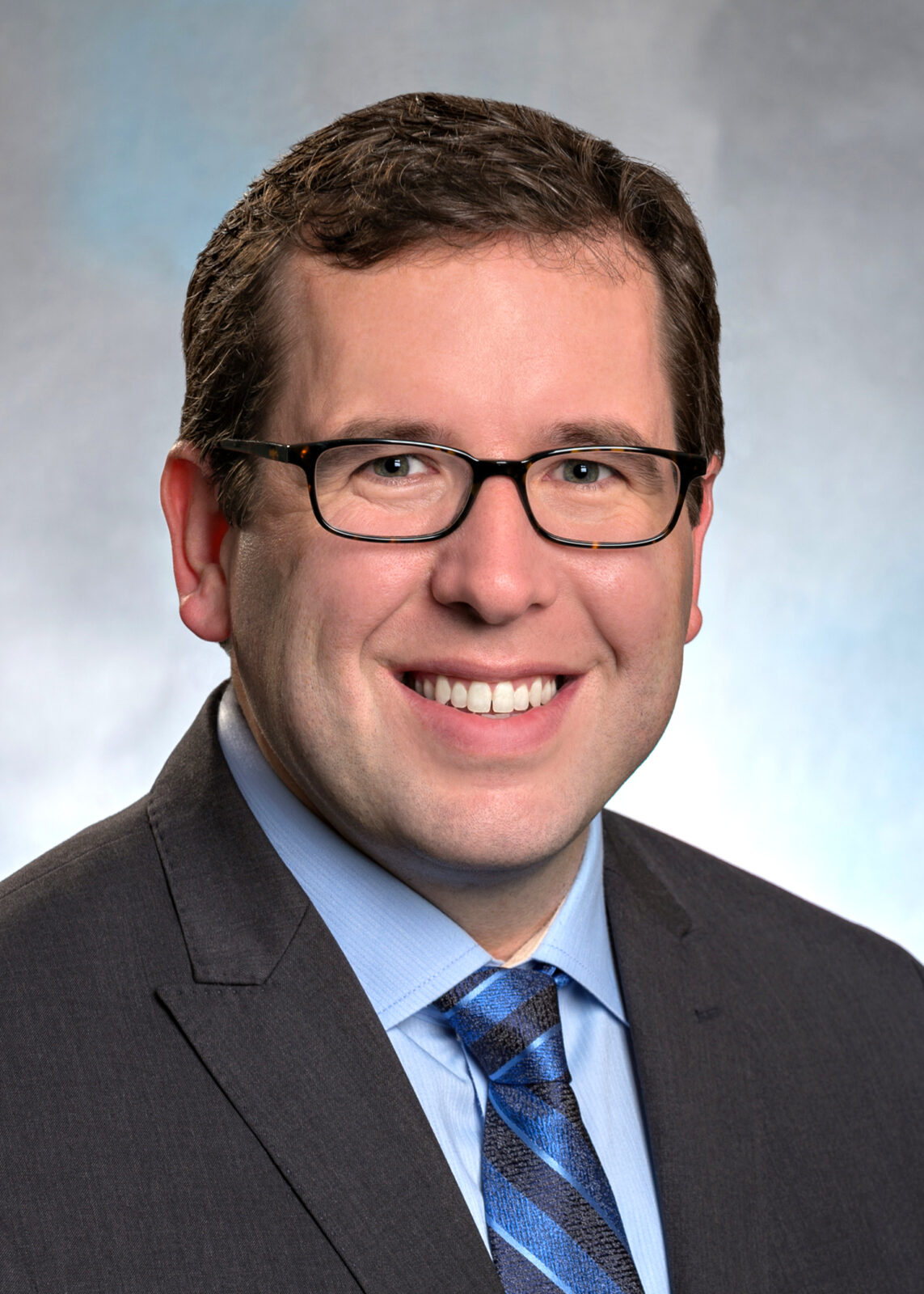 July 2024, man with dark hair and glasses with grey jacket, blue shirt and tie against a light blue background